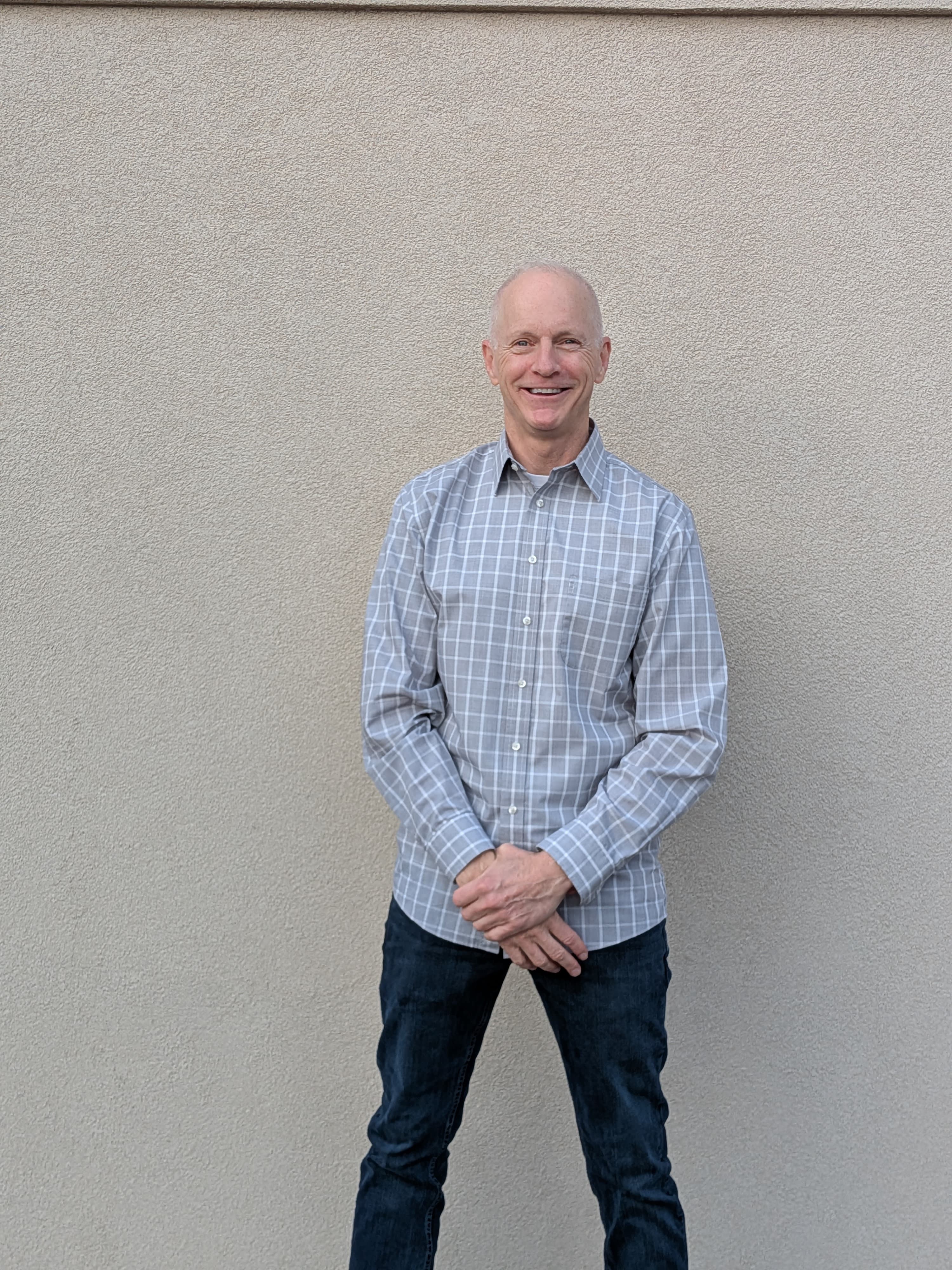 Smiling man wearing a grey plaid shirt and dark jeans stands against a textured wall.