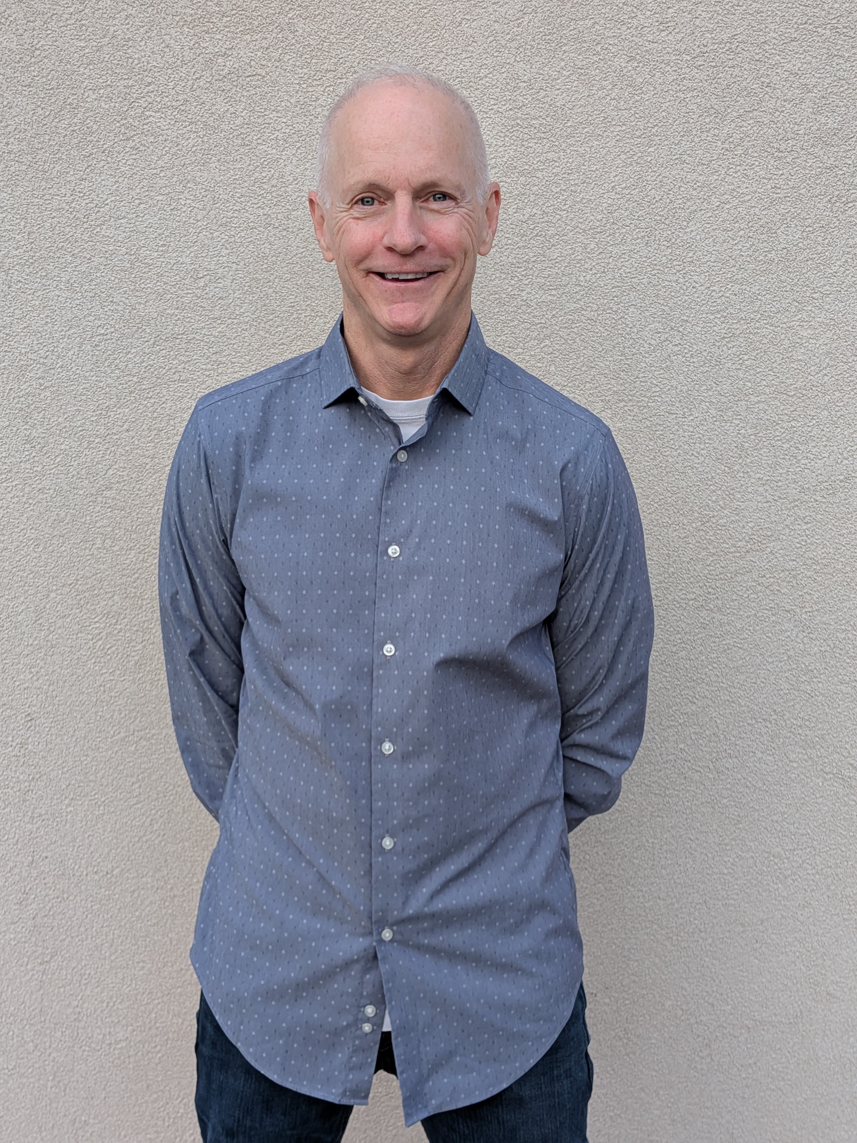 Smiling man in a blue patterned shirt stands with hands behind back against textured wall.