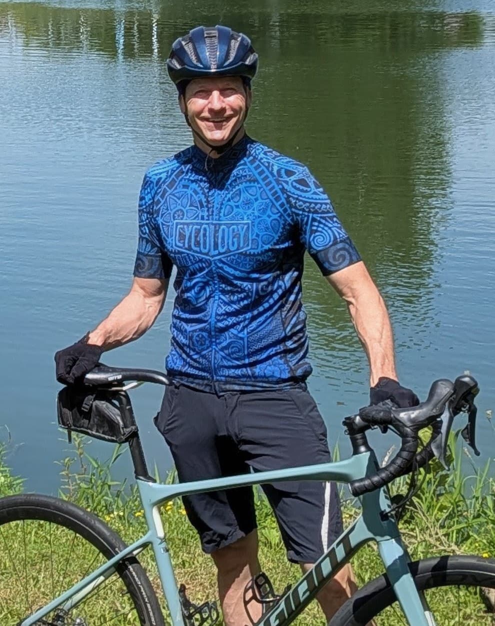 Smiling man in blue patterned cycling jersey stands with a road bike by a lake.